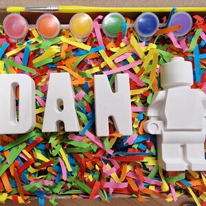 May include: A craft kit with white ceramic letters spelling "DAN" and a white ceramic figurine of a person. The kit includes paint pots in various colors and a paintbrush. The letters and figurine are surrounded by colorful paper shreds.
