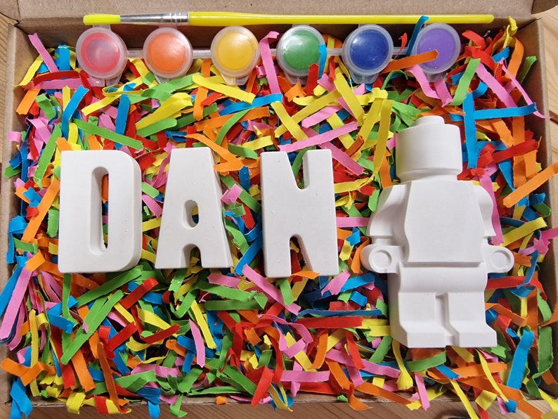 May include: A craft kit with white ceramic letters spelling "DAN" and a white ceramic figurine of a person. The kit includes paint pots in various colors and a paintbrush. The letters and figurine are surrounded by colorful paper shreds.