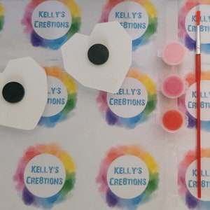 May include: Two white ceramic heart-shaped ornaments with black magnets on the back. The ornaments are surrounded by rainbow-colored circles with the text "Kelly's Creations" in white. Three small jars of paint in pink, red, and orange are included, along with a red paintbrush.