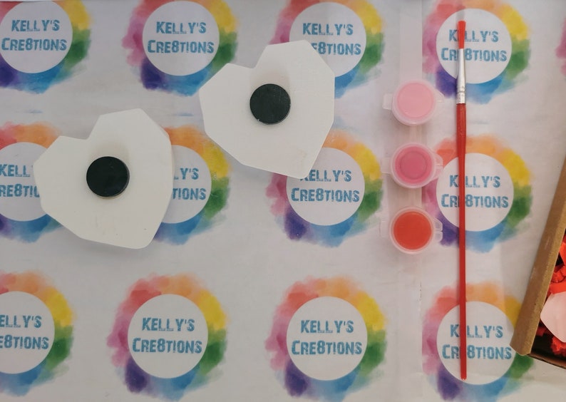 May include: Two white ceramic heart-shaped ornaments with black magnets on the back. The ornaments are surrounded by rainbow-colored circles with the text "Kelly's Creations" in white. Three small jars of paint in pink, red, and orange are included, along with a red paintbrush.