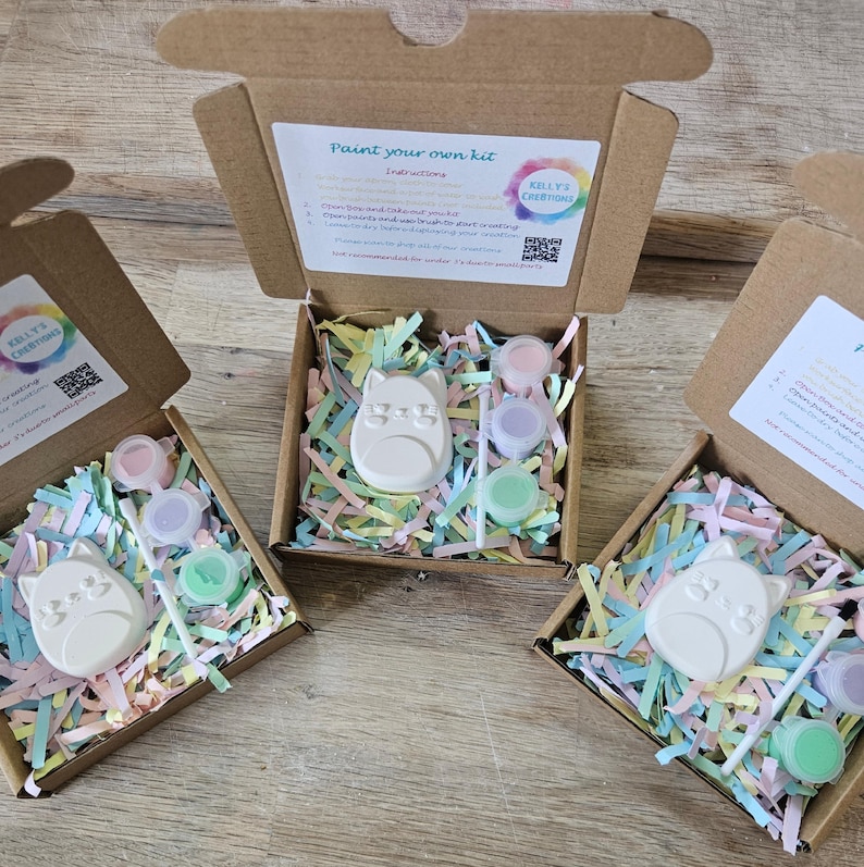 Puede incluir: Tres cajas de cart&oacute;n abiertas que contienen un kit de pintura. Cada kit incluye una figura de gato de cer&aacute;mica blanca, un pincel peque&ntilde;o y tres botes de pintura circulares en rosa, morado y verde, sobre tiras de papel de colores pastel.