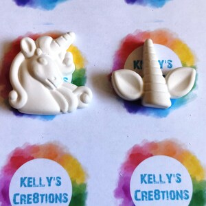 Puede incluir: Dos piezas artesanales blancas con tem&aacute;tica de unicornio. Una es una figura de unicornio completa, la otra es un cuerno de unicornio con orejas. Las piezas est&aacute;n sobre una superficie blanca con detalles de arco&iacute;ris y el texto "Kelly's Creations".