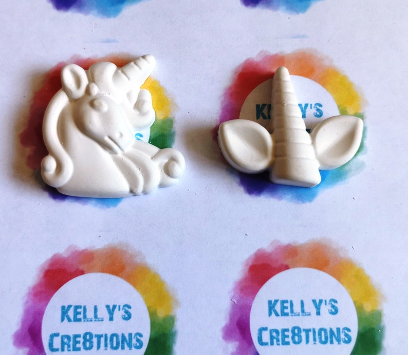 Puede incluir: Dos piezas artesanales blancas con tem&aacute;tica de unicornio. Una es una figura de unicornio completa, la otra es un cuerno de unicornio con orejas. Las piezas est&aacute;n sobre una superficie blanca con detalles de arco&iacute;ris y el texto "Kelly's Creations".