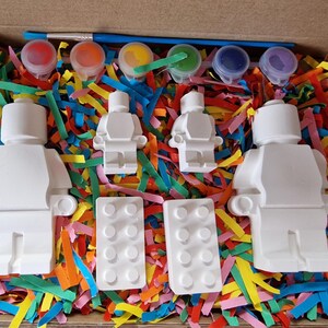 May include: A paint-your-own Lego minifigure kit with six paint pots, a paintbrush, and two Lego bricks. The kit is in a cardboard box with colorful shredded paper.