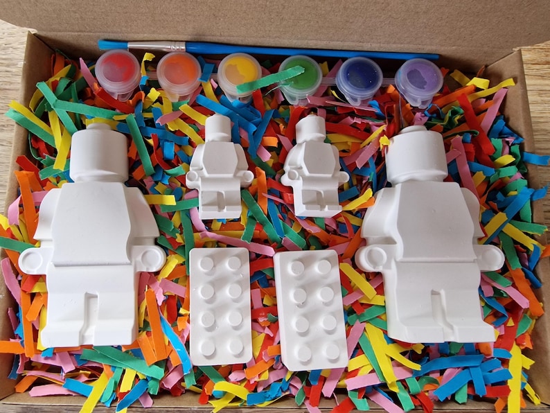 May include: A paint-your-own Lego minifigure kit with six paint pots, a paintbrush, and two Lego bricks. The kit is in a cardboard box with colorful shredded paper.