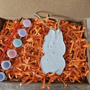 May include: A craft kit in a cardboard box, featuring a white snowman-shaped ornament with a jute string, surrounded by orange paper shreds. The kit includes small paint pots in various colors and a red paintbrush.