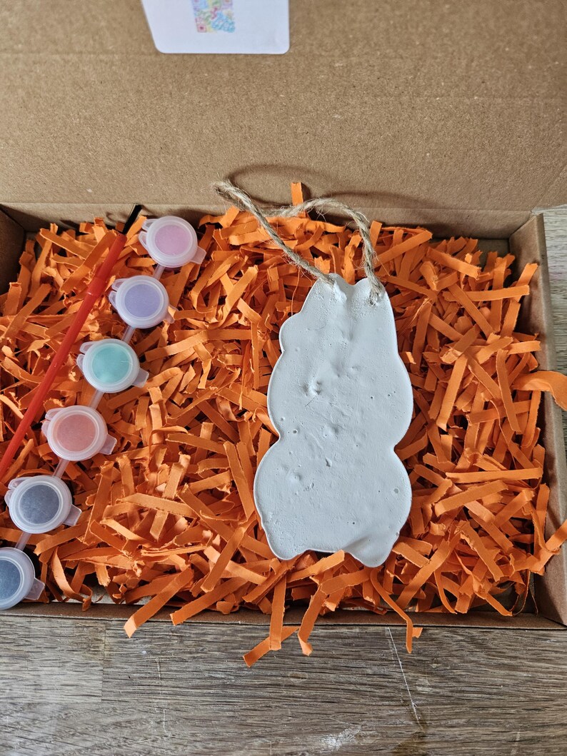 May include: A craft kit in a cardboard box, featuring a white snowman-shaped ornament with a jute string, surrounded by orange paper shreds. The kit includes small paint pots in various colors and a red paintbrush.