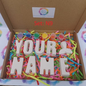 May include: A craft kit in a cardboard box, featuring the words "YOUR NAME" in large, white block letters, a mermaid tail, and small paint pots. The box is filled with colorful paper shreds. The text "Set: N9" is visible.