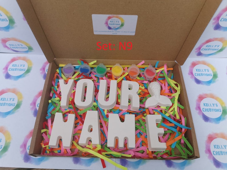 May include: A craft kit in a cardboard box, featuring the words "YOUR NAME" in large, white block letters, a mermaid tail, and small paint pots. The box is filled with colorful paper shreds. The text "Set: N9" is visible.
