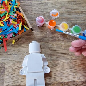 May include: A white plastic figurine of a Lego person, ready to be painted.  The figurine is on a wooden surface with a set of paint pots and a paintbrush.