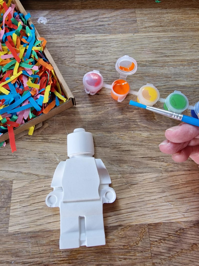 May include: A white plastic figurine of a Lego person, ready to be painted.  The figurine is on a wooden surface with a set of paint pots and a paintbrush.