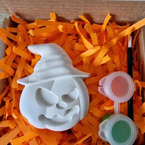 May include: A white, unpainted pumpkin-shaped craft kit with a witch hat, nestled in orange paper strips inside a cardboard box. Includes two paint pots, one red and one green, and a small paintbrush. The text "Not recommended for under 3's due to small parts" is visible.