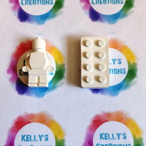 Puede incluir: Dos figuritas de hormig&oacute;n blanco en miniatura de un personaje Lego y un ladrillo Lego. Las figuritas est&aacute;n sobre una superficie blanca con c&iacute;rculos de colores.