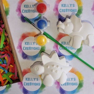 May include: A craft kit with two white ceramic dinosaur figurines, a paintbrush, and four small paint pots with red, blue, yellow, and green paint. The kit is packaged in a white box with the text "Kelly's Creations" printed on it.