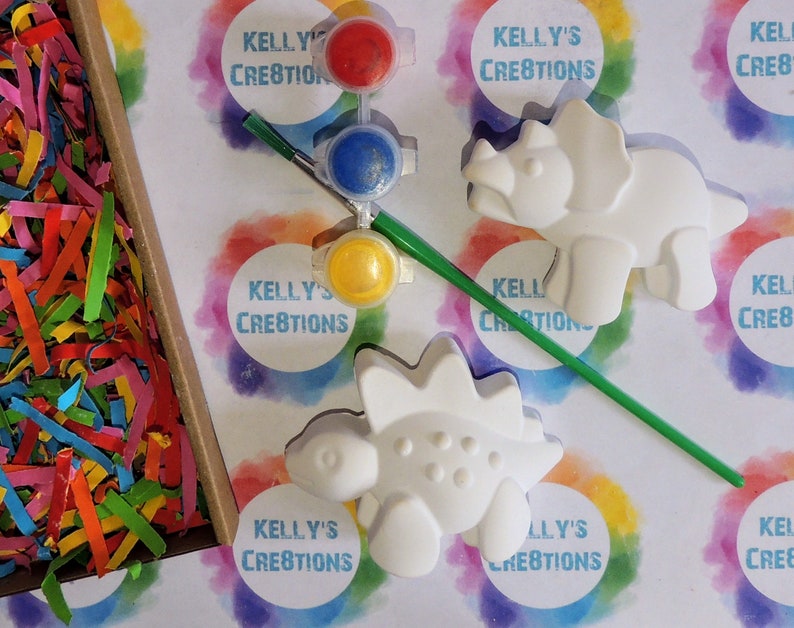 May include: A craft kit with two white ceramic dinosaur figurines, a paintbrush, and four small paint pots with red, blue, yellow, and green paint. The kit is packaged in a white box with the text "Kelly's Creations" printed on it.