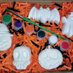 May include: A Halloween-themed paint-your-own craft kit with five white ceramic figures: a pumpkin, a ghost, a bat, a skull, and a witch's hat. The kit includes six small pots of paint in various colors and a paintbrush.
