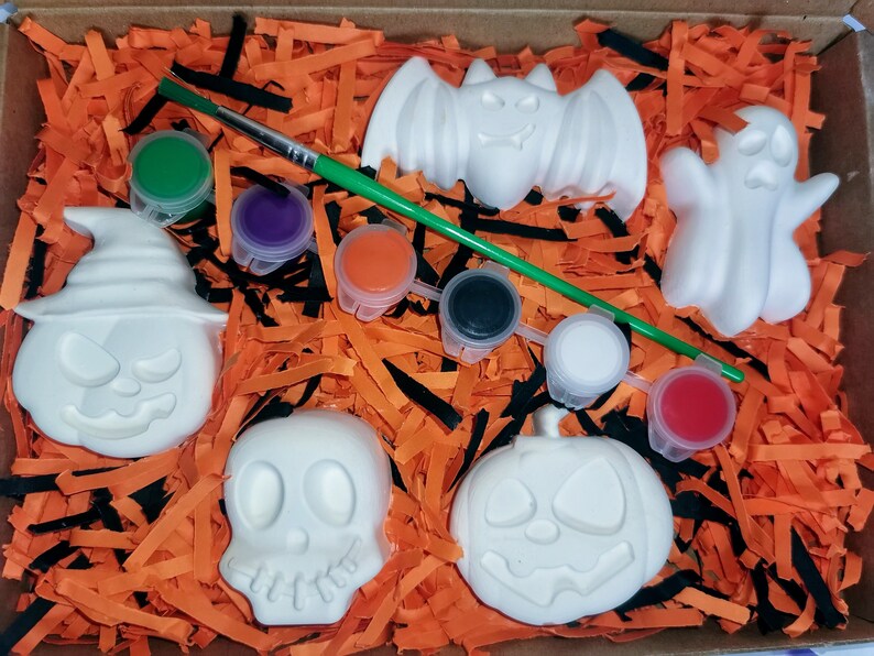 May include: A Halloween-themed paint-your-own craft kit with five white ceramic figures: a pumpkin, a ghost, a bat, a skull, and a witch's hat. The kit includes six small pots of paint in various colors and a paintbrush.