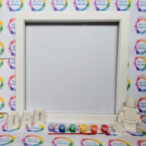 May include: A white paint-by-numbers kit featuring a white Lego figure and the word "DAD" in white letters. The kit includes a white frame, a paintbrush, and six small paint pots in various colors.
