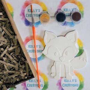 May include: A white ceramic fox figurine with paint-by-numbers outlines. The kit includes three small paint pots with lids, a paintbrush, and a sheet of paper with the Kelly's Creations logo.