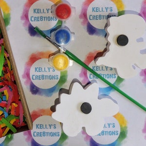 May include: Two white wooden flower shapes with magnets on the back, paint pots in red, blue, and yellow, a paintbrush, and a bag of colorful shredded paper. The paint pots and the wooden shapes have the text "Kelly's Creations" on them.