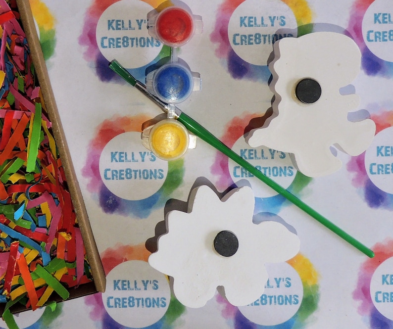 May include: Two white wooden flower shapes with magnets on the back, paint pots in red, blue, and yellow, a paintbrush, and a bag of colorful shredded paper. The paint pots and the wooden shapes have the text "Kelly's Creations" on them.