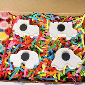 May include: A set of four white ceramic cars with black circles in the middle, ready to be painted. The cars are in a box with colorful shredded paper and a set of paint pots.