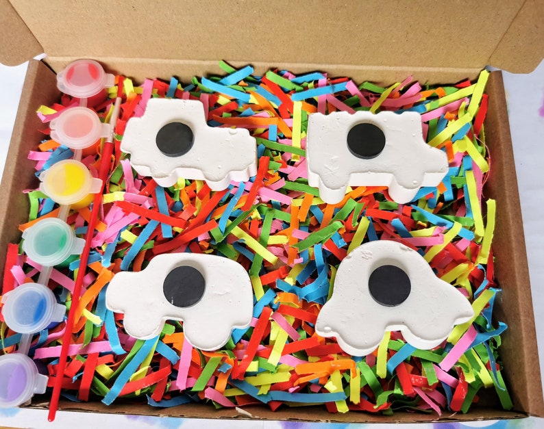 May include: A set of four white ceramic cars with black circles in the middle, ready to be painted. The cars are in a box with colorful shredded paper and a set of paint pots.