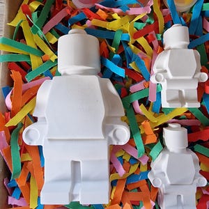 May include: Three white ceramic figurines of Lego people, in different sizes, in a box with colorful shredded paper and paint pots.