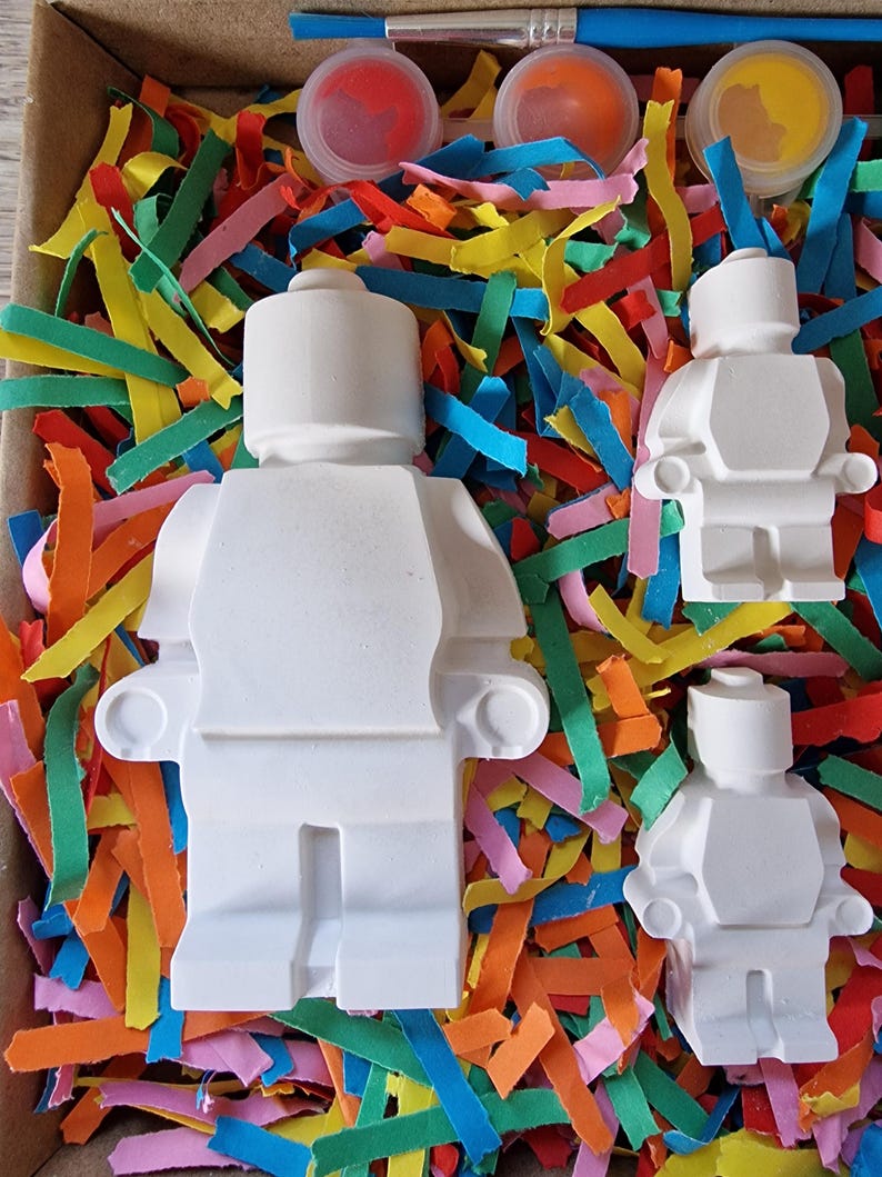 May include: Three white ceramic figurines of Lego people, in different sizes, in a box with colorful shredded paper and paint pots.