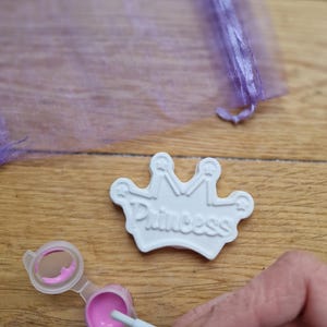 May include: White ceramic crown with the word "Princess" embossed on it. The crown is being painted with pink paint.