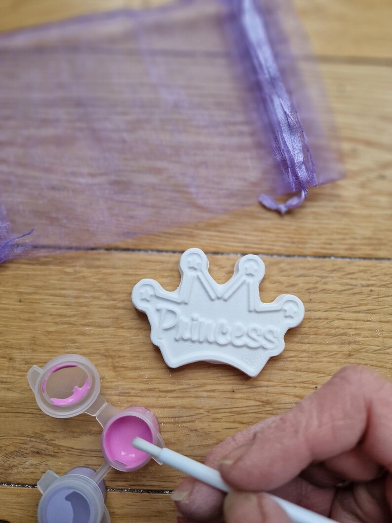May include: White ceramic crown with the word "Princess" embossed on it. The crown is being painted with pink paint.