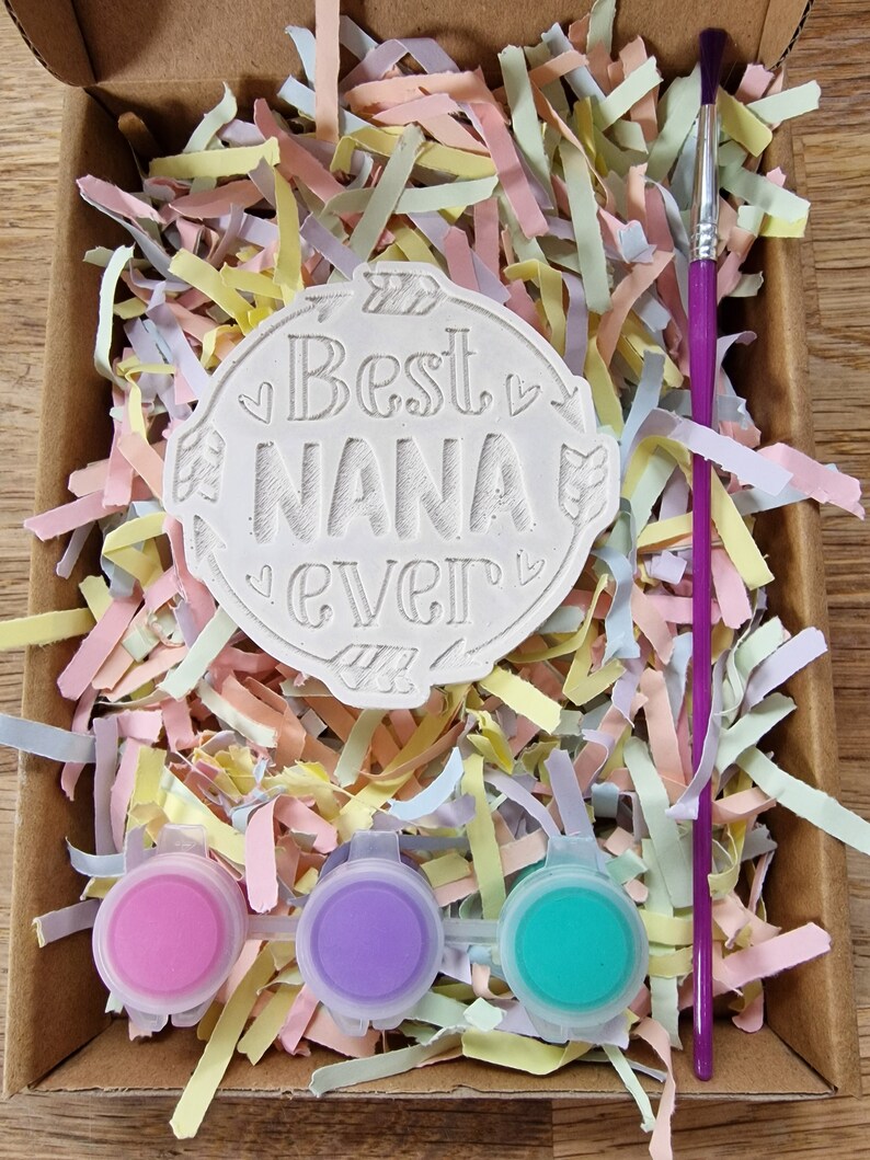 May include: A craft kit with a white ceramic tile that says "Best Nana Ever" in a decorative font. The kit includes three small bottles of paint in pink, purple, and turquoise, and a purple paintbrush.