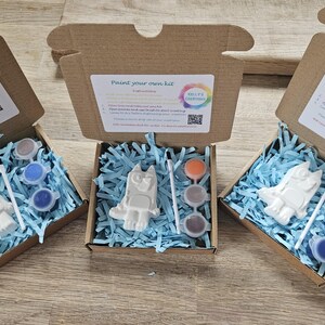 May include: Three open cardboard boxes, each containing a paint-your-own kit. Each kit includes a white figurine, paint pots in various colours, and a paintbrush, all nestled in blue shredded paper. The boxes are labelled "Paint your own kit."