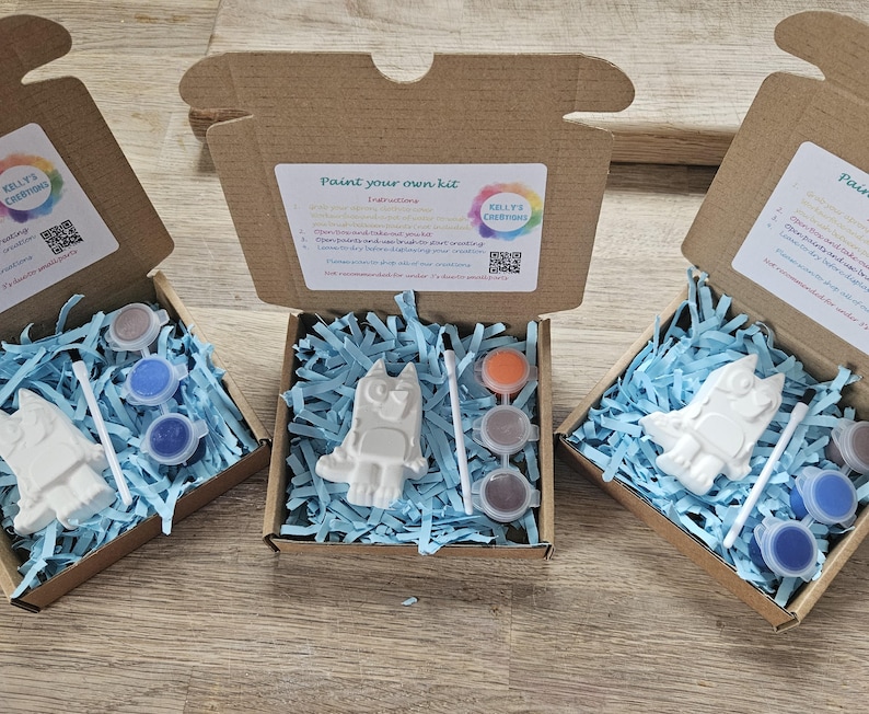 May include: Three open cardboard boxes, each containing a paint-your-own kit. Each kit includes a white figurine, paint pots in various colours, and a paintbrush, all nestled in blue shredded paper. The boxes are labelled "Paint your own kit."