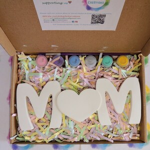Pu&ograve; includere: Un kit per lavoretti per dipingere un cartello "MAMMA". Il kit include lettere in ceramica bianca, un cuore e sei piccoli flaconi di vernice. Le lettere e il cuore sono circondati da carta triturata colorata.