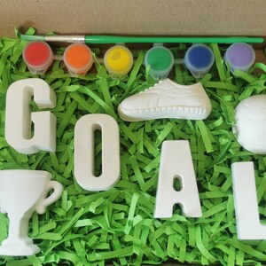 May include: A craft kit with white ceramic letters spelling "GOAL", a soccer ball, a shoe, a trophy, and six paint pots with a paintbrush. The items are arranged on a bed of green shredded paper.