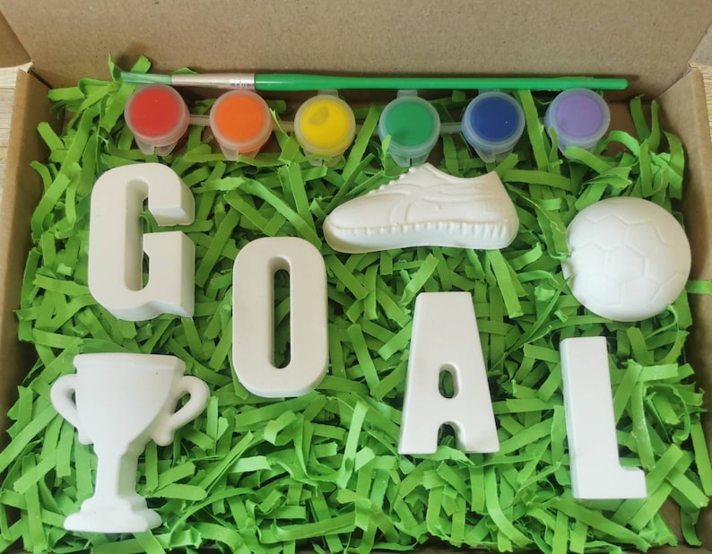 May include: A craft kit with white ceramic letters spelling "GOAL", a soccer ball, a shoe, a trophy, and six paint pots with a paintbrush. The items are arranged on a bed of green shredded paper.