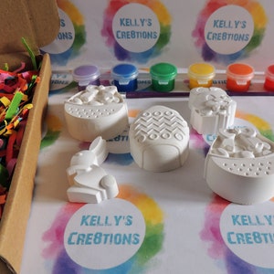 May include: A set of five white ceramic Easter-themed figurines, including a bunny, an egg, and a basket, ready to be painted. The set comes with six small jars of paint in various colors, including blue, green, yellow, and red.