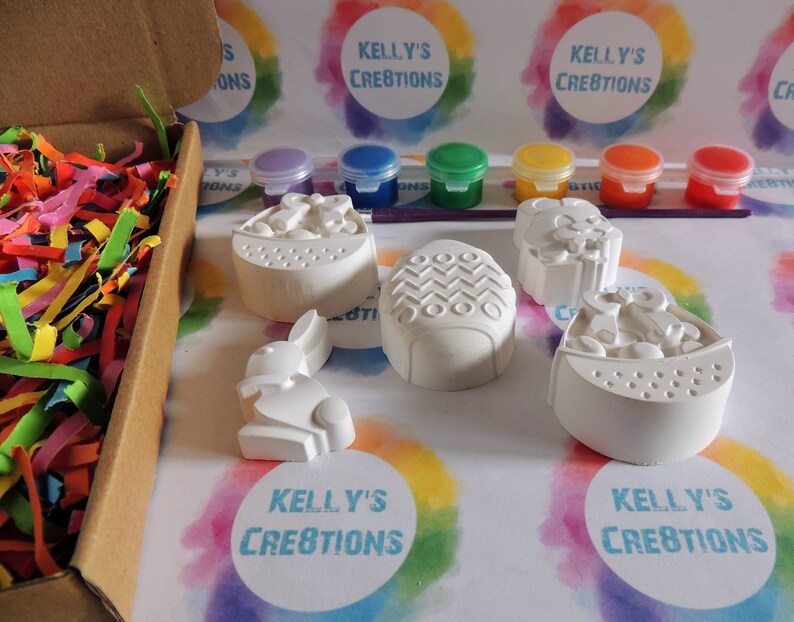 May include: A set of five white ceramic Easter-themed figurines, including a bunny, an egg, and a basket, ready to be painted. The set comes with six small jars of paint in various colors, including blue, green, yellow, and red.