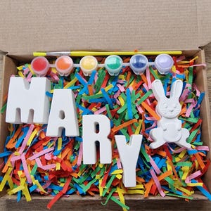 May include: A craft kit with white ceramic letters spelling "MARY" and a white ceramic bunny. The kit includes five small paint pots in various colors, a paintbrush, and a yellow stick. The letters and bunny are sitting on a bed of colorful shredded paper.