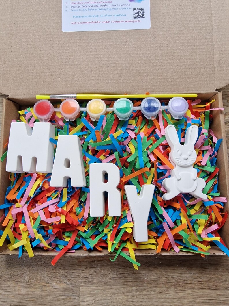 May include: A craft kit with white ceramic letters spelling "MARY" and a white ceramic bunny. The kit includes five small paint pots in various colors, a paintbrush, and a yellow stick. The letters and bunny are sitting on a bed of colorful shredded paper.