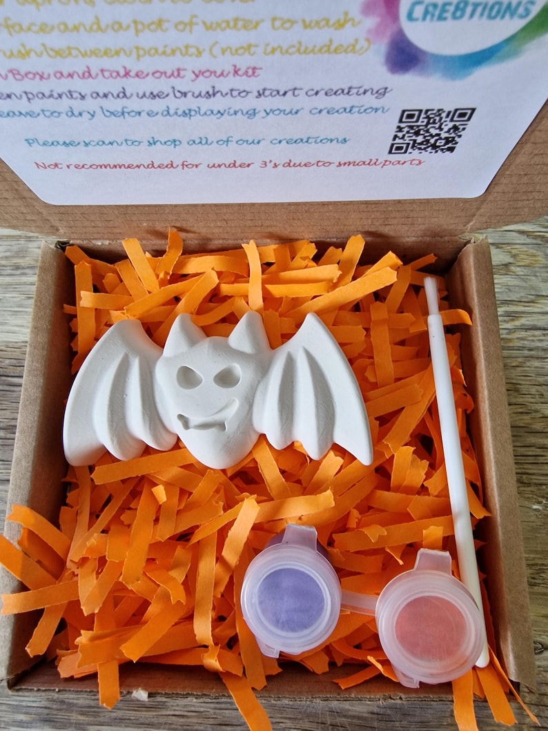 May include: A craft kit featuring a white bat figurine, paint pots, and a paintbrush, nestled in orange paper shreds inside a cardboard box. The kit includes instructions and a QR code for more creations.