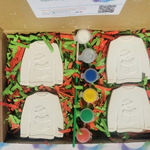 May include: A set of four white ceramic ornaments shaped like sweaters with a snowman design. The ornaments are packaged in a brown box with green and red shredded paper. The box also includes four bottles of paint and a paintbrush.