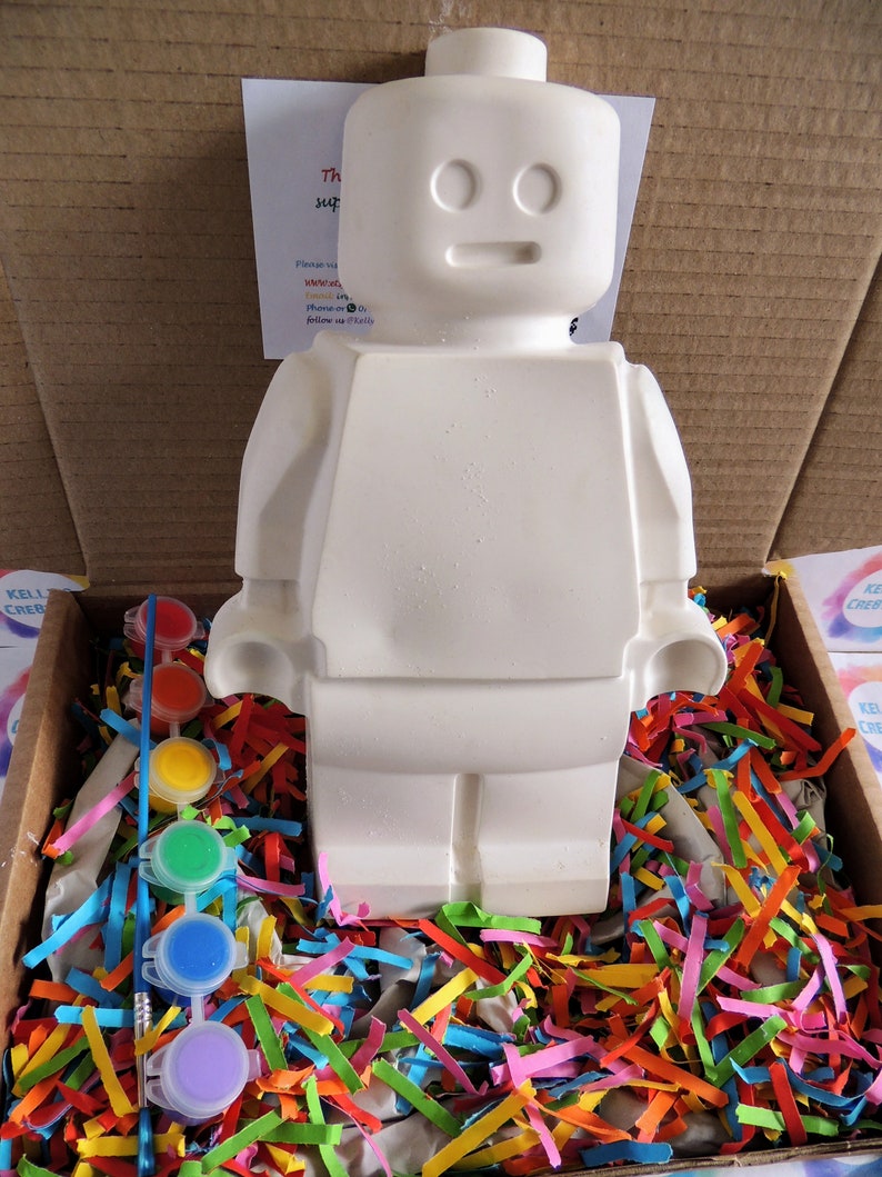 May include: A white ceramic figurine of a Lego person, with a paintbrush and six small paint pots in various colors, all sitting in a box filled with colorful shredded paper.
