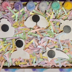 Puede incluir: Un conjunto de cinco adornos de cer&aacute;mica blanca en forma de nube, arco&iacute;ris, unicornio, sirena y estrella. Cada adorno tiene un c&iacute;rculo negro en el centro. Los adornos est&aacute;n rodeados de papel triturado de colores en una caja de cart&oacute;n. La caja est&aacute; etiquetada con "Kelly's Creations".