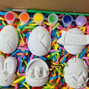 May include: A set of six white ceramic Easter-themed ornaments in a box with colourful shredded paper. The ornaments include a bunny, a basket, an egg with the words "Happy Easter", and three other eggs with different designs. The box also includes six small bottles of paint.