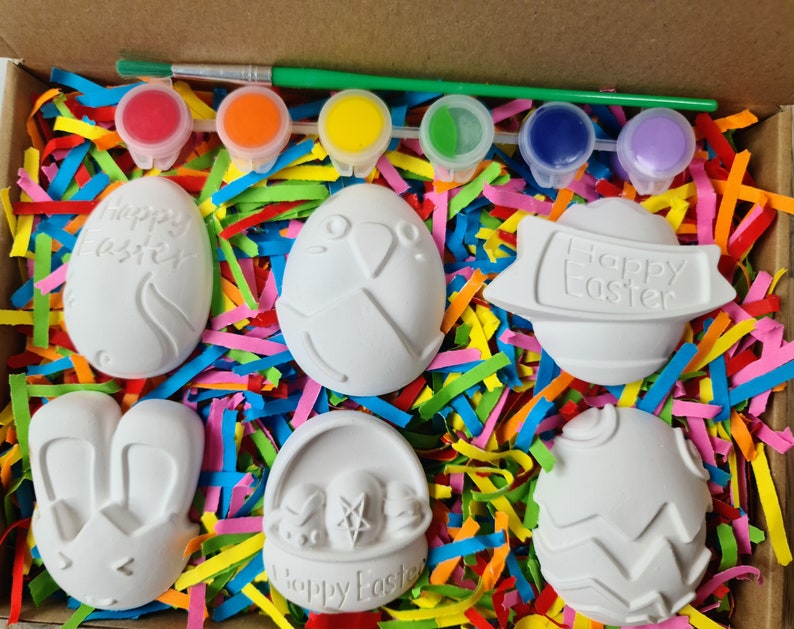 May include: A set of six white ceramic Easter-themed ornaments in a box with colourful shredded paper. The ornaments include a bunny, a basket, an egg with the words "Happy Easter", and three other eggs with different designs. The box also includes six small bottles of paint.