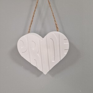 Puede incluir: Un letrero blanco en forma de coraz&oacute;n con la palabra "PRIDE" en relieve en la parte delantera. El letrero est&aacute; suspendido por un cord&oacute;n de hilo natural y cuelga de una pared gris. El letrero es un art&iacute;culo decorativo.