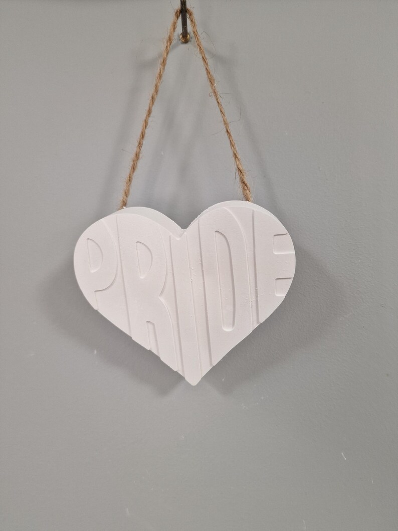 Puede incluir: Un letrero blanco en forma de coraz&oacute;n con la palabra "PRIDE" en relieve en la parte delantera. El letrero est&aacute; suspendido por un cord&oacute;n de hilo natural y cuelga de una pared gris. El letrero es un art&iacute;culo decorativo.
