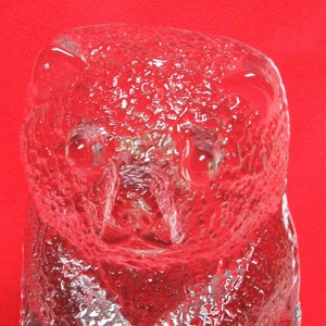 May include: A clear glass sculpture of a bear. The bear is sitting with its paws crossed and its head tilted down. The glass is textured and has a frosted appearance.